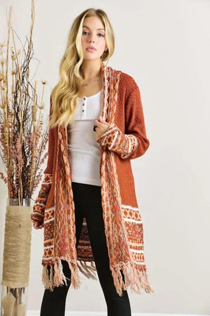 Woman wearing a rust orange knit cardigan with fringe, white top, and black pants