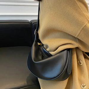 Black leather shoulder bag with gold zipper worn over beige coat on black sofa