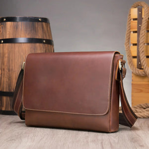 Brown leather messenger bag with adjustable strap, displayed indoors on wooden floor