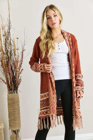 Woman wearing rust orange patterned knit cardigan with fringe over white top and black pants