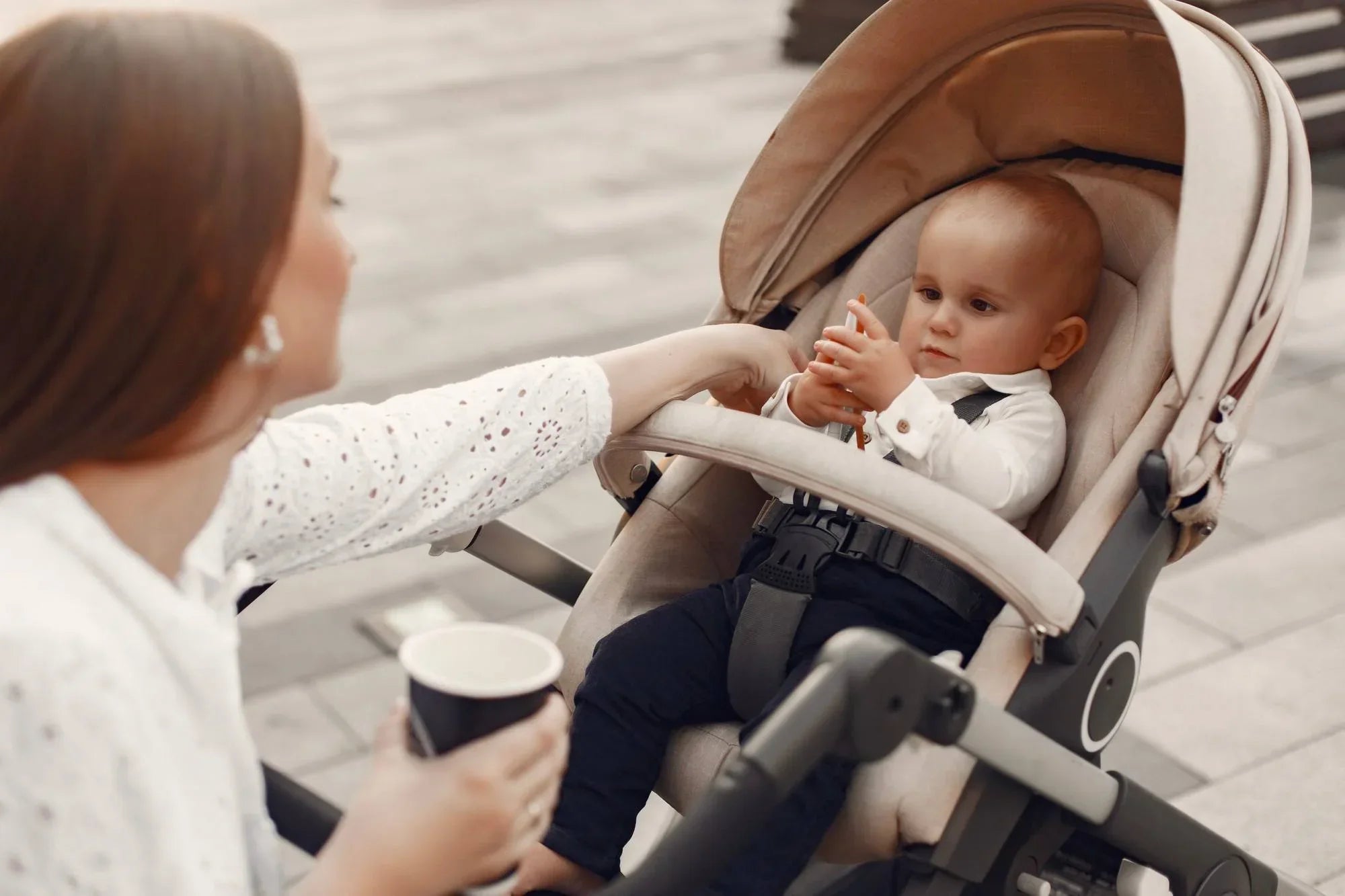 Woman with coffee cup interacts with baby in beige stroller on city sidewalk