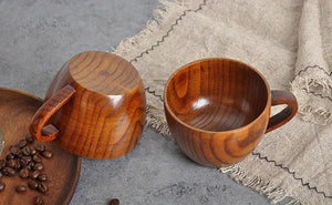 Two wooden coffee cups with handles on a textured gray surface, next to coffee beans and linen fabric.