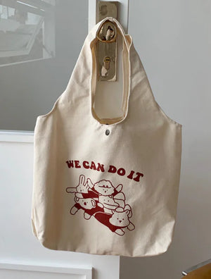 Beige canvas tote bag with cartoon animal print and 'We Can Do It' slogan, hanging on door.