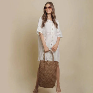 Woman in white embroidered dress holding large woven straw tote bag, wearing sunglasses, beige background