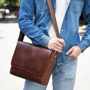 Man wearing blue denim shirt and jeans, carrying brown leather messenger bag outdoors