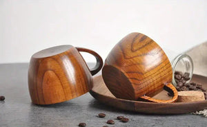 Two wooden coffee cups with handles on a table, coffee beans and a wooden tray nearby