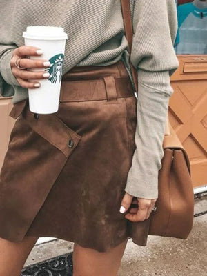 Woman wearing khaki sweater and brown suede skirt, holding coffee and tan leather shoulder bag