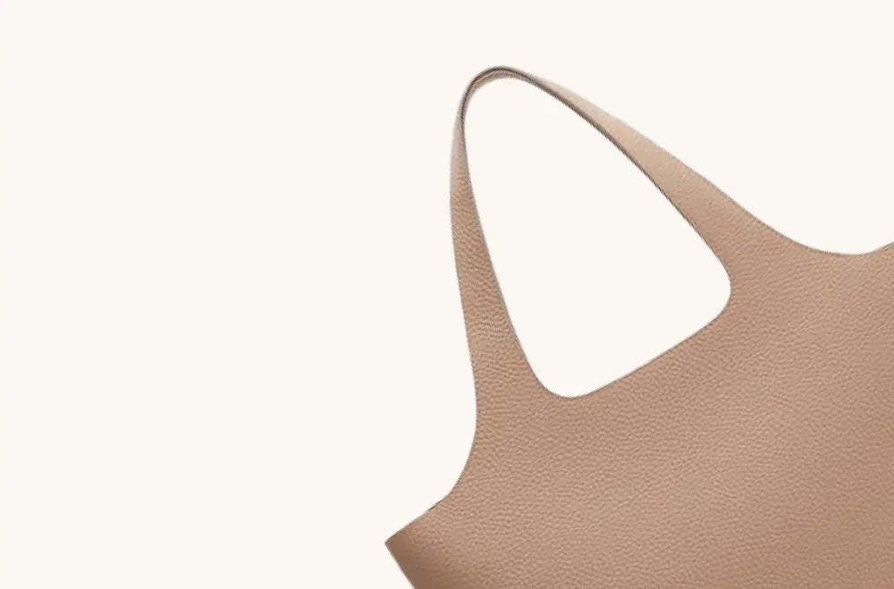 Beige leather tote bag with textured finish and soft curved handles on neutral background