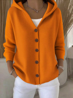 Woman wearing a bright orange button-up knit hoodie with white pants and layered jewelry