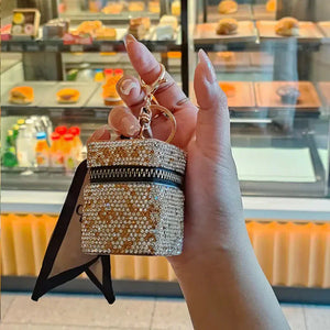 Hand holding a small rhinestone-studded jewelry box keychain in front of a bakery display