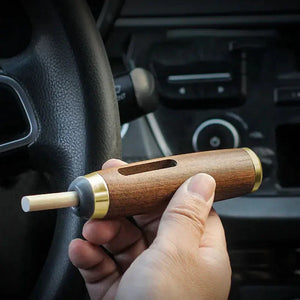 Hand holding a wooden car air freshener with gold accents near a steering wheel
