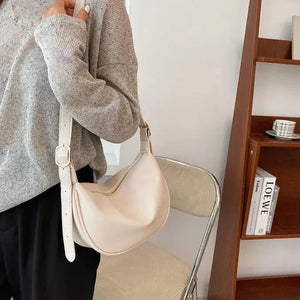 Woman wearing a light gray sweater and black pants, holding a cream shoulder bag indoors