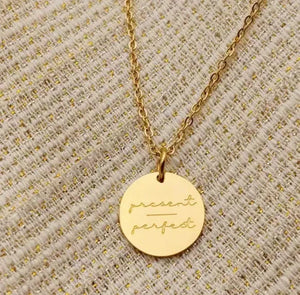 Gold necklace with round pendant engraved 'present perfect' on textured fabric background