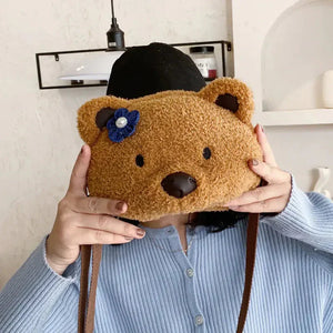 Plush teddy bear face shoulder bag with blue flower, held by person in light blue top