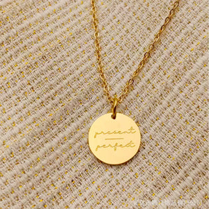 Gold round pendant necklace engraved with 'present perfect' on textured fabric background